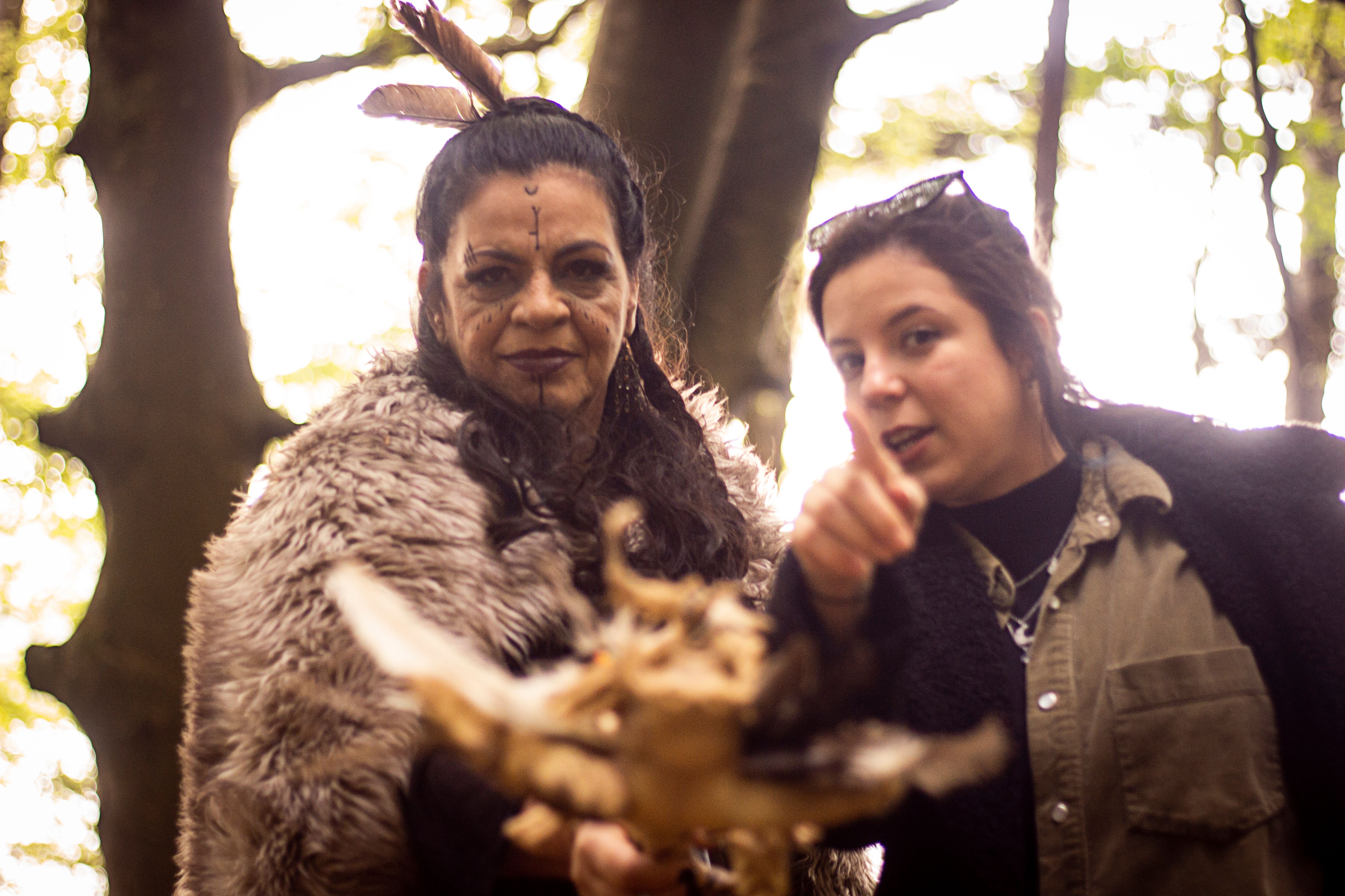 two female figures look towards viewer - one is clad in furs holding a large staff with feagthers in their hair